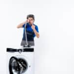 Repairman in uniform pointing at a pipe behind a washing machine, illustrating Whirlpool washer error code FP drain fault