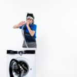 Repairman with a head lamp clearing a pipe behind a washing machine, illustrating Whirlpool washer FP drain fault error code
