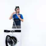 Confused repairman in uniform holding a plastic pipe behind a washing machine, illustrating Whirlpool washer F13 error code dispenser fault