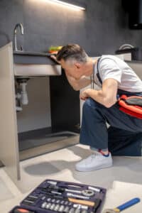 Technician kneeling to inspect pipes under the sink for a jammed garbage disposal