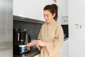 Woman in a bathrobe placing a blue cup under a black countertop coffee machine in a home kitchen