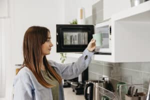 Woman placing a mug inside an open countertop microwave in a white kitchen