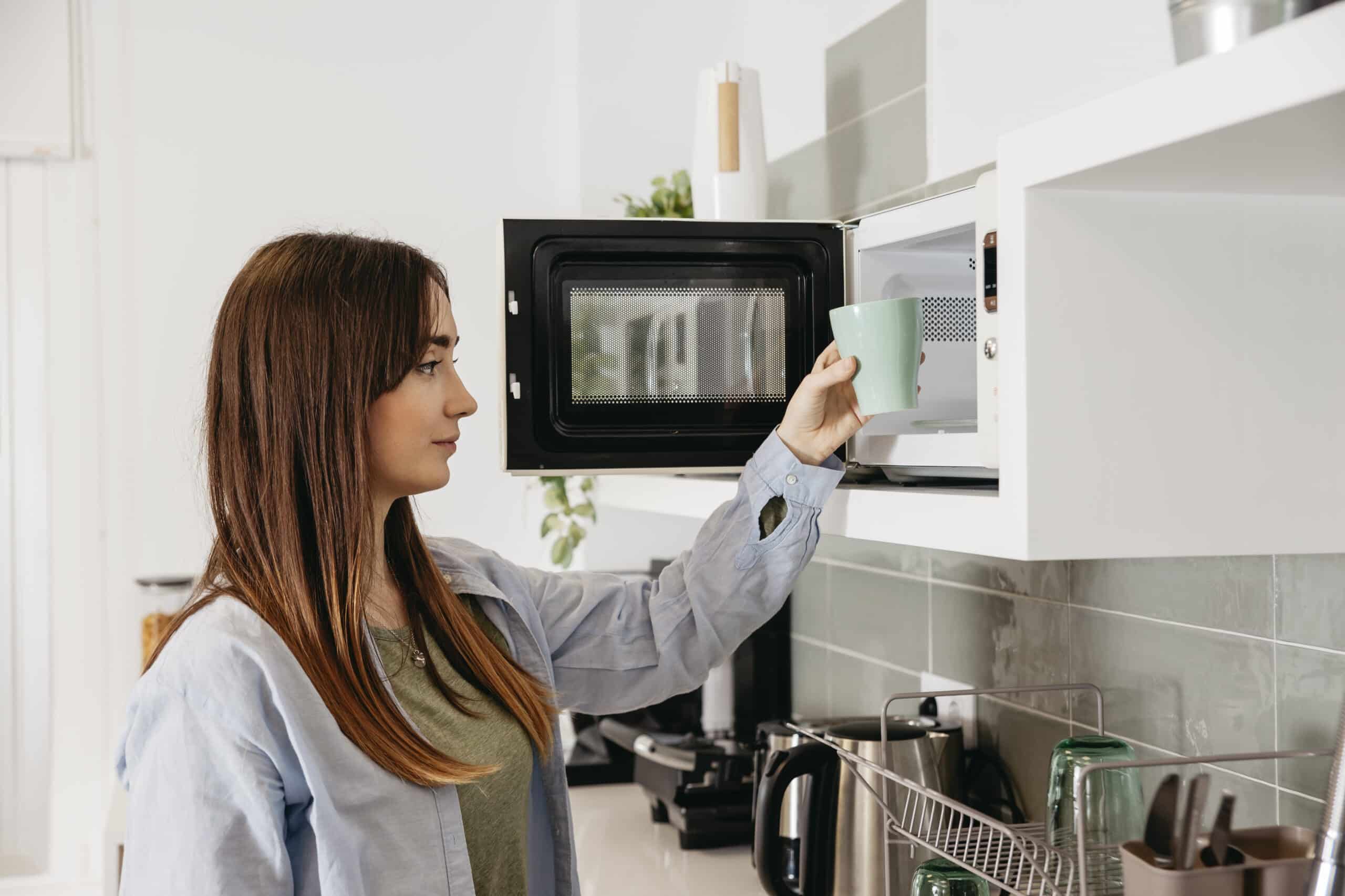Woman placing a mug inside an open countertop microwave in a white kitchen