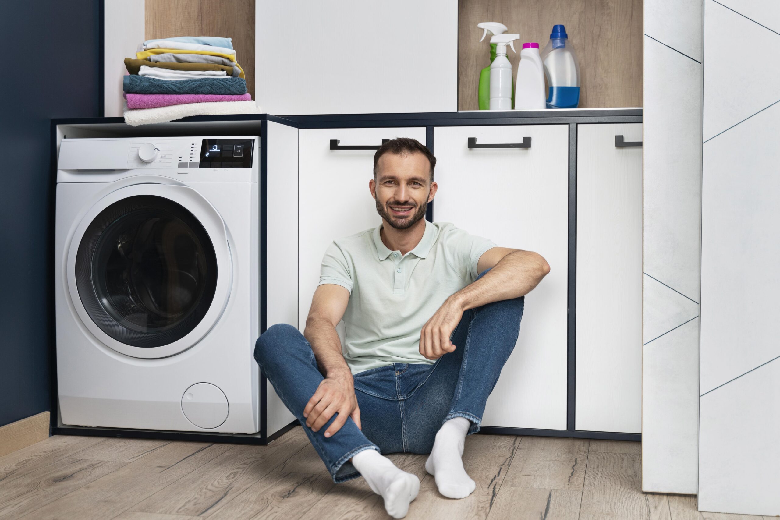 Man sitting on the floor next to a closed front-load washing machine, waiting for the cycle to finish