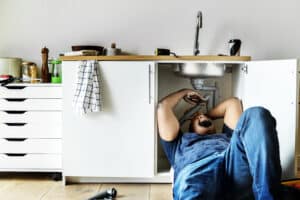 Man lying on a kitchen floor, reaching up to work on the drain pipes inside the open cabinet under the sink