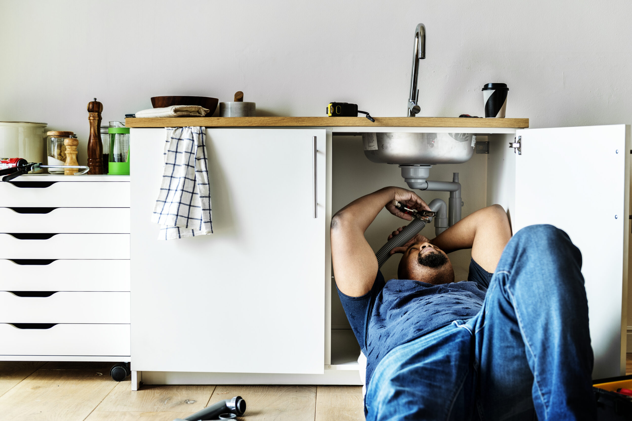 Man lying on a kitchen floor, reaching up to work on the drain pipes inside the open cabinet under the sink