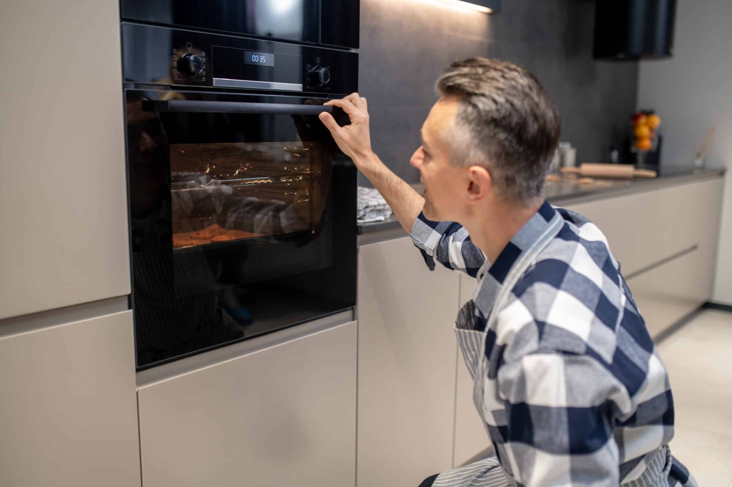 Man kneeling at a built-in oven, turning a control knob while food bakes inside